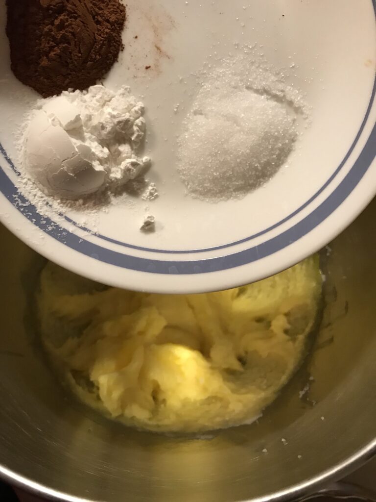 A small plate holding ground cinnamon, baking powder, and salt being held over a mixing bowl containing creamed butter and sugar.
