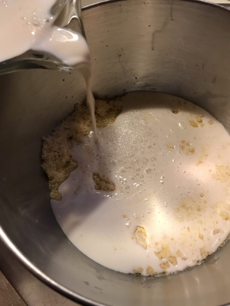 Milk being poured from a glass pitcher into a stainless steel mixing bowl containing a crumbly flour and butter mixture.