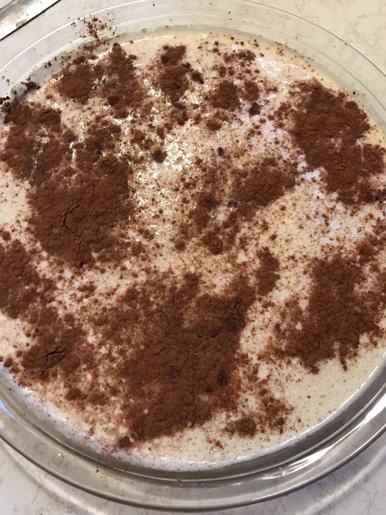 Milk tart batter in a glass dish generously topped with a dusting of ground cinnamon before being placed in the oven.