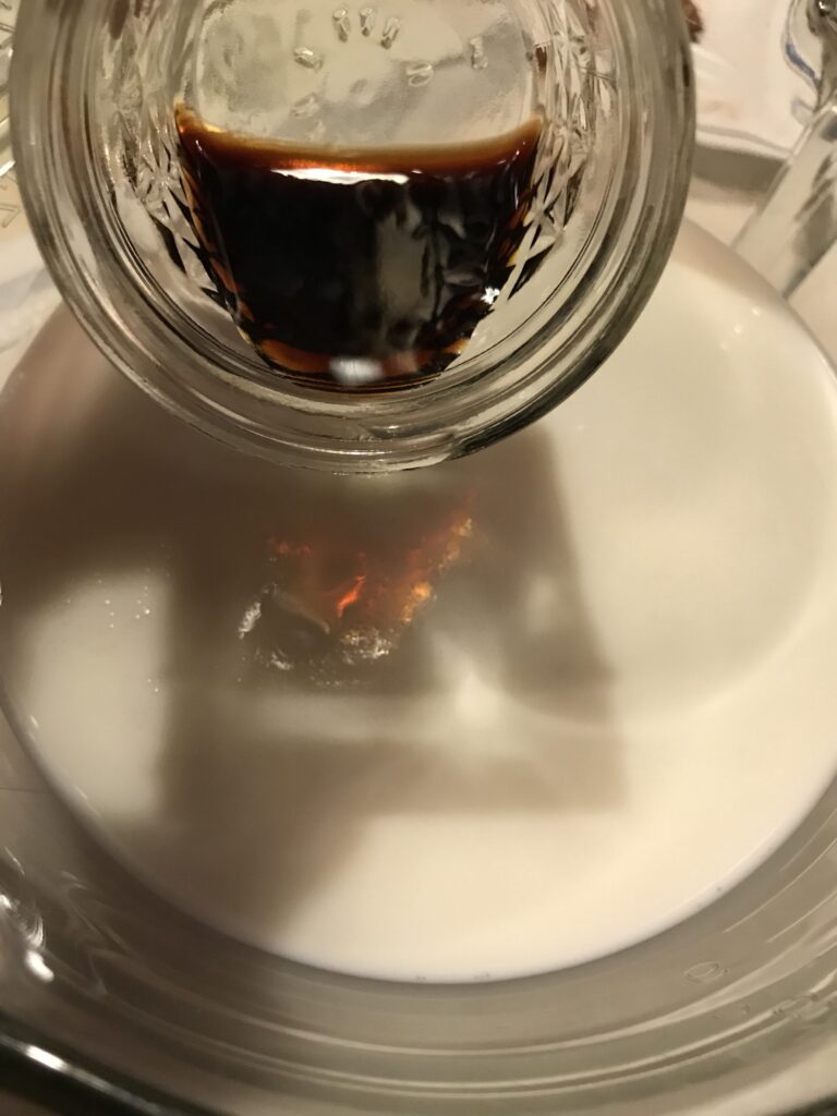 Dark vanilla extract being poured from a small glass mason jar into a large bowl of white milk for a tart filling.