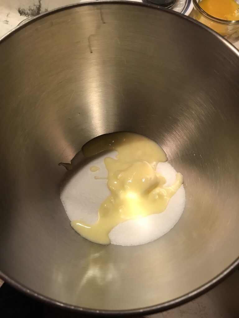 Softened butter being added to white granulated sugar in a large stainless steel mixing bowl for a milk tart recipe.
