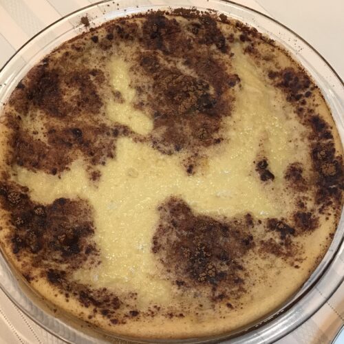 A fully baked milk tart in a glass dish with a golden-brown cinnamon-dusted crust, resting on a striped tablecloth.