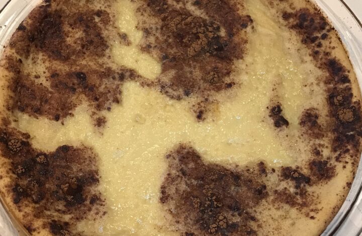 A fully baked milk tart in a glass dish with a golden-brown cinnamon-dusted crust, resting on a striped tablecloth.