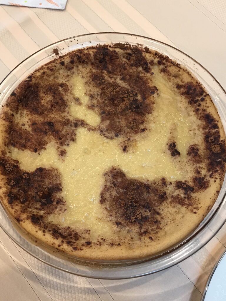 A fully baked milk tart in a glass dish with a golden-brown cinnamon-dusted crust, resting on a striped tablecloth.