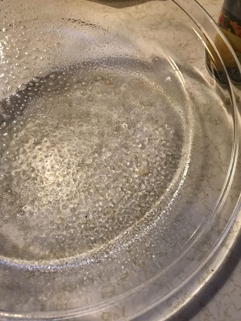 A clear glass round pie dish sitting on a white countertop, thoroughly greased with butter or oil to prevent the milk tart from sticking.