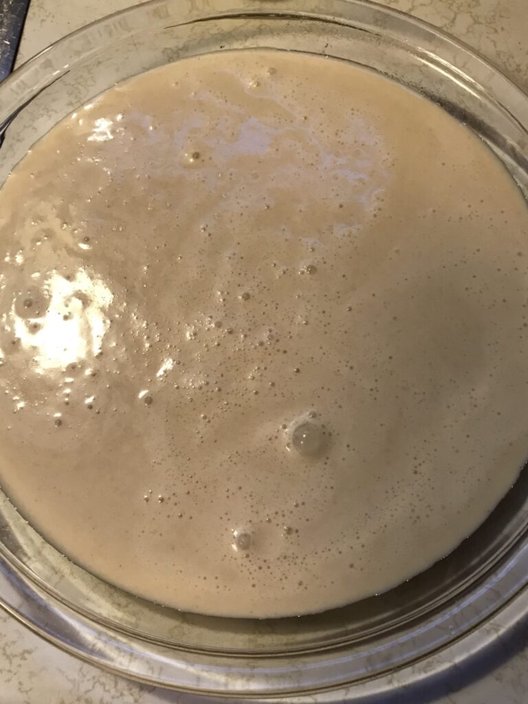 Milk tart batter with small surface bubbles poured into a round, greased glass baking dish on a white countertop.