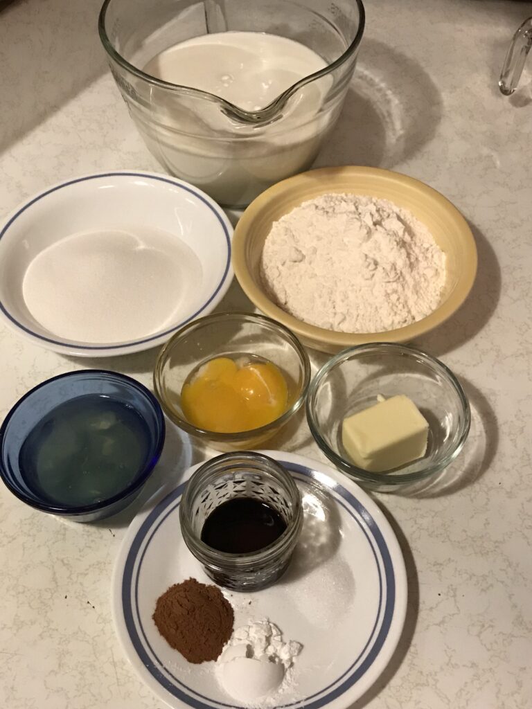 A bird's-eye view of ingredients for a traditional milk tart, including bowls of flour, sugar, eggs, butter, milk, and cinnamon spices on a white counter.