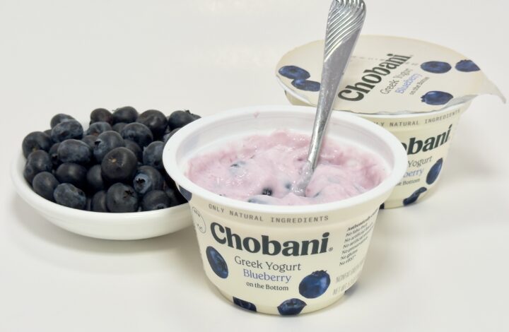 A mixed cup of Chobani Blueberry Greek yogurt with a spoon, shown next to the foil lid and a white bowl overflowing with fresh blueberries.