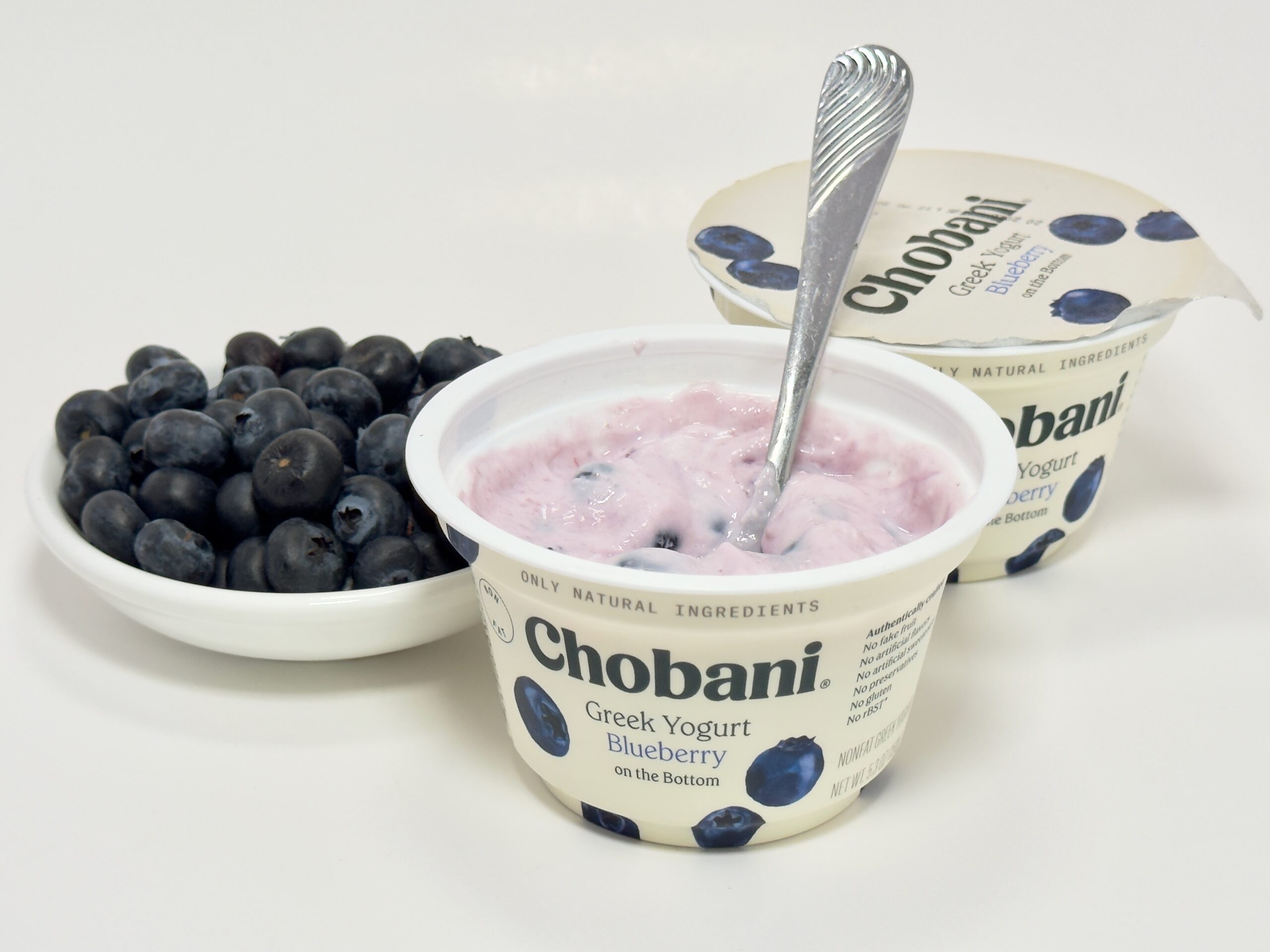 A mixed cup of Chobani Blueberry Greek yogurt with a spoon, shown next to the foil lid and a white bowl overflowing with fresh blueberries.