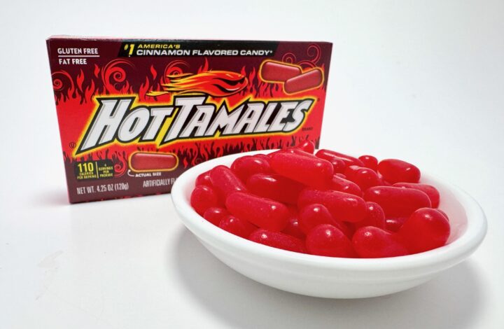 A 4.25 oz box of Hot Tamales Fierce Cinnamon candy standing behind a white bowl overflowing with the red chewy candies.