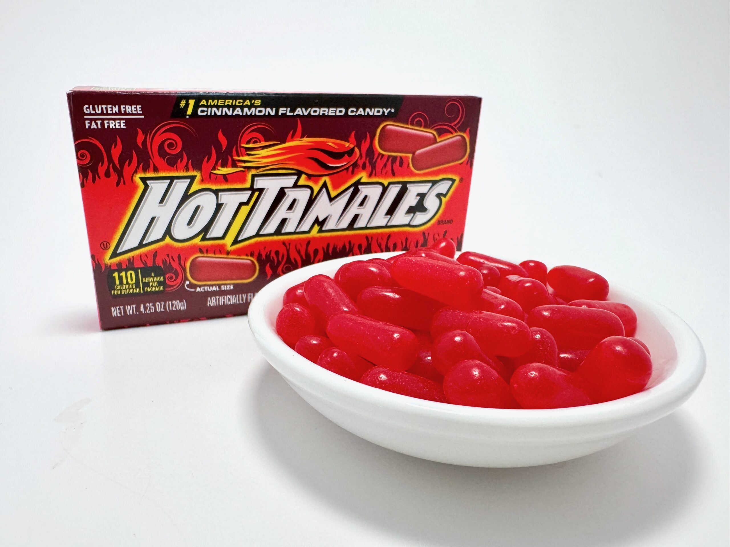 A 4.25 oz box of Hot Tamales Fierce Cinnamon candy standing behind a white bowl overflowing with the red chewy candies.
