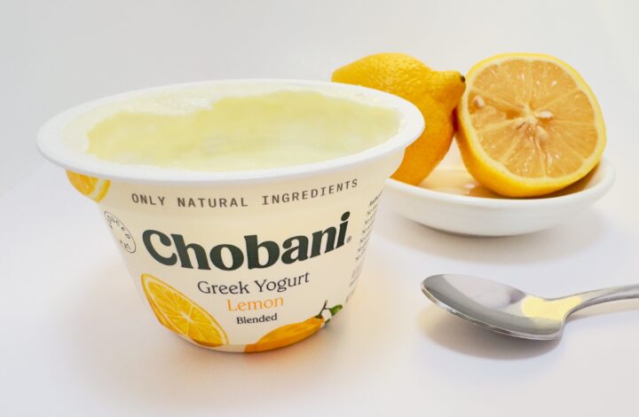 Chobani Greek Lemon Blended yogurt cup next to a sliced whole, fresh lemon and spoon on a white background.