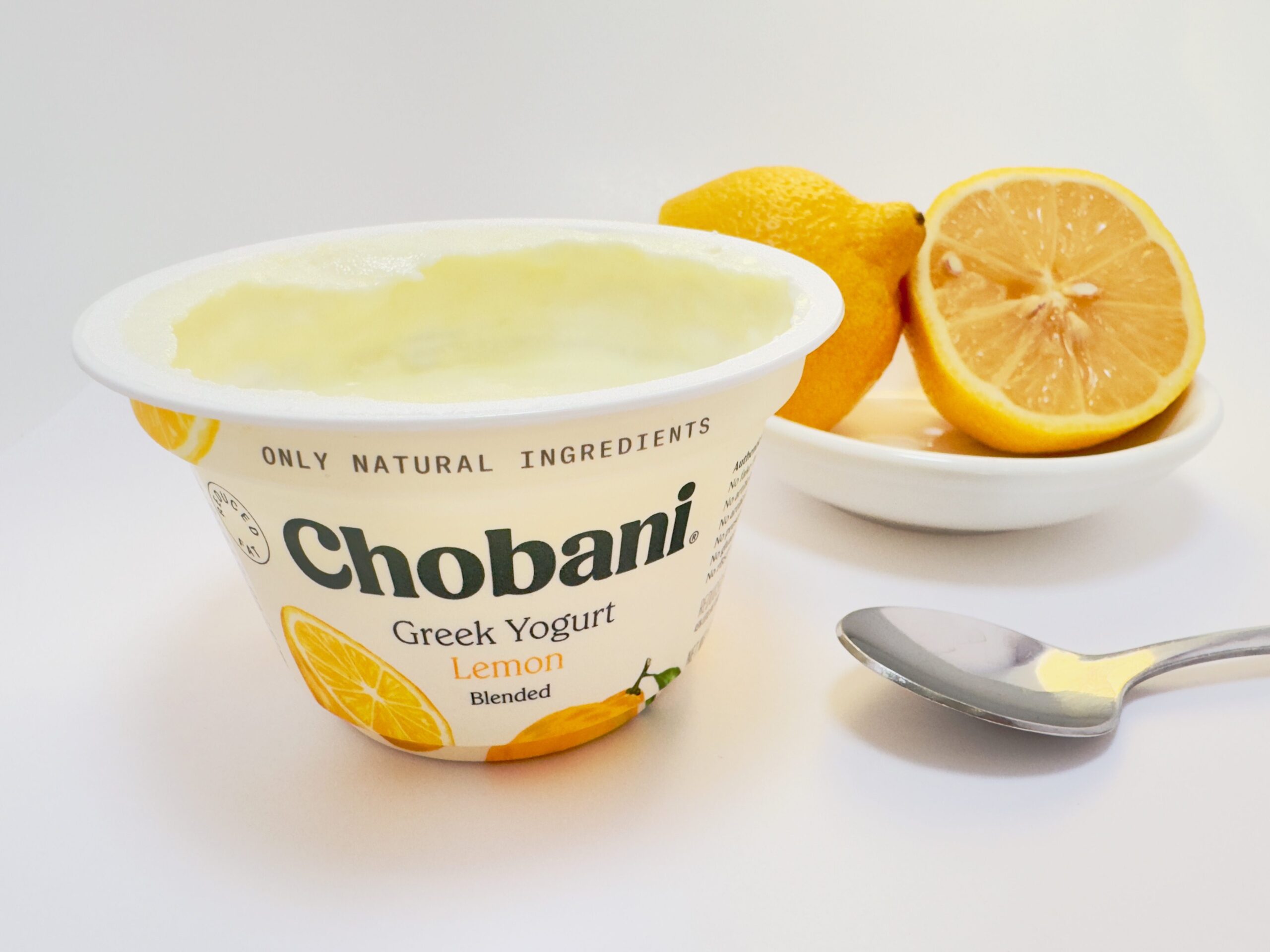 Chobani Greek Lemon Blended yogurt cup next to a sliced whole, fresh lemon and spoon on a white background.