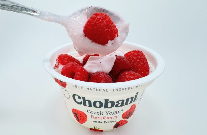 A spoonful of light pink Chobani Raspberry Greek yogurt topped with a large, fresh red raspberry, held over a yogurt cup filled with more fresh berries.
