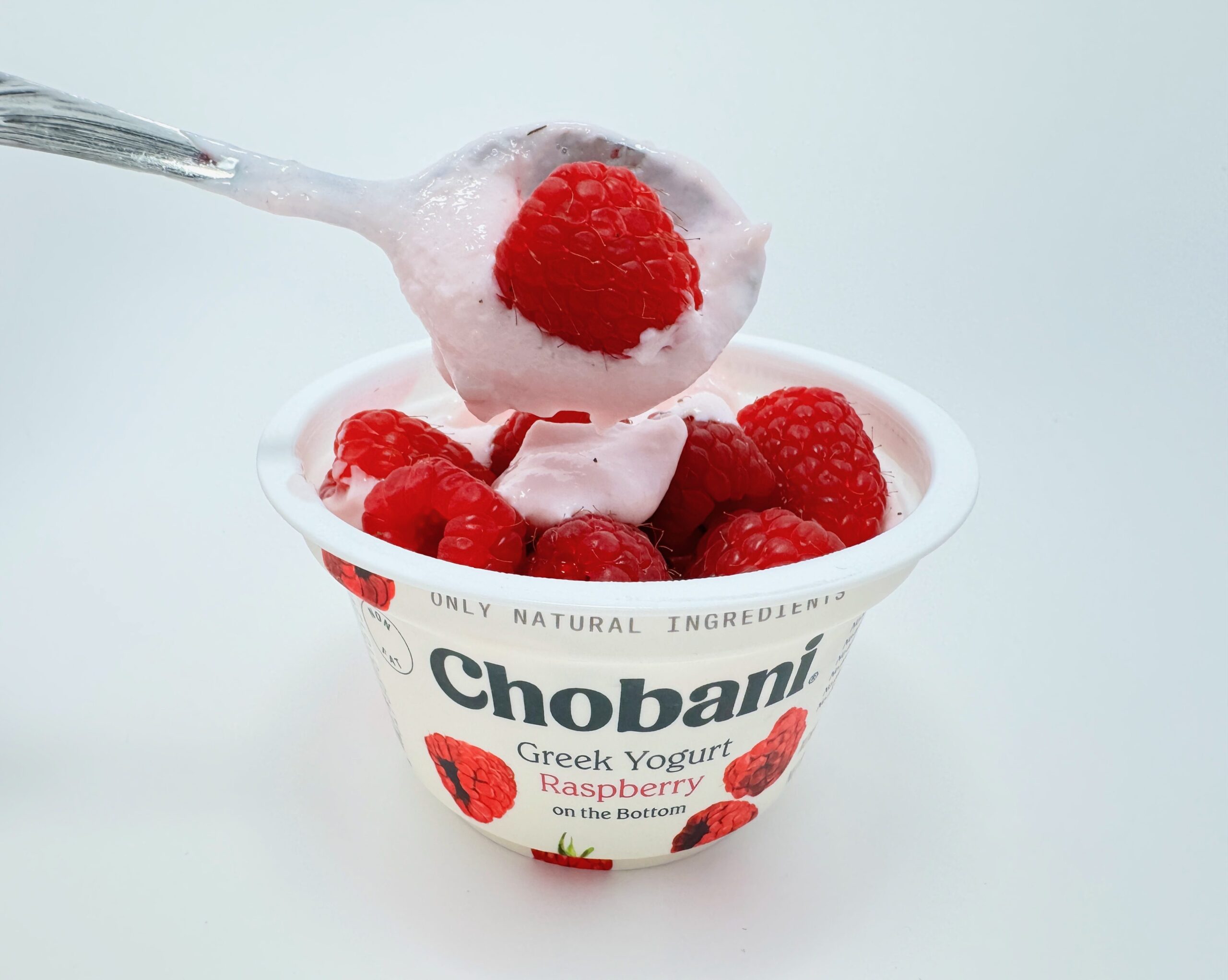 A spoonful of light pink Chobani Raspberry Greek yogurt topped with a large, fresh red raspberry, held over a yogurt cup filled with more fresh berries.