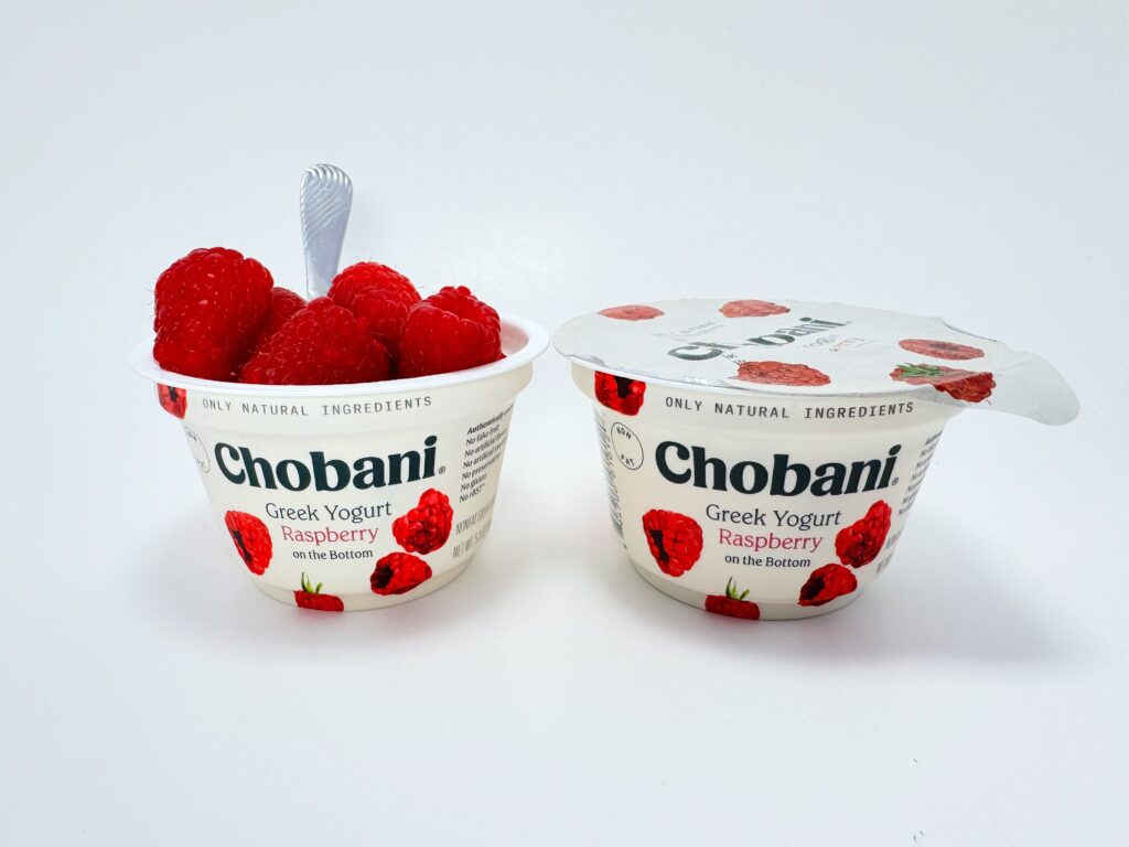 Two cups of Chobani Greek Yogurt Raspberry on the Bottom: one is closed, and the other is open and filled with fresh raspberries and a spoon. Used for a product comparison in a food review.