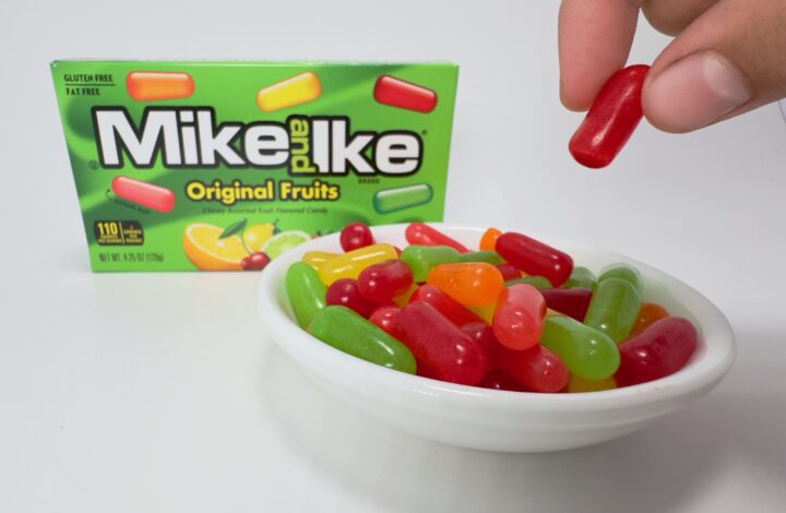 Hand picking up a green Mike and Ike Original Fruits candy from a bowl of mixed cherry, lemon, lime, strawberry, and orange candies with the box in the background.