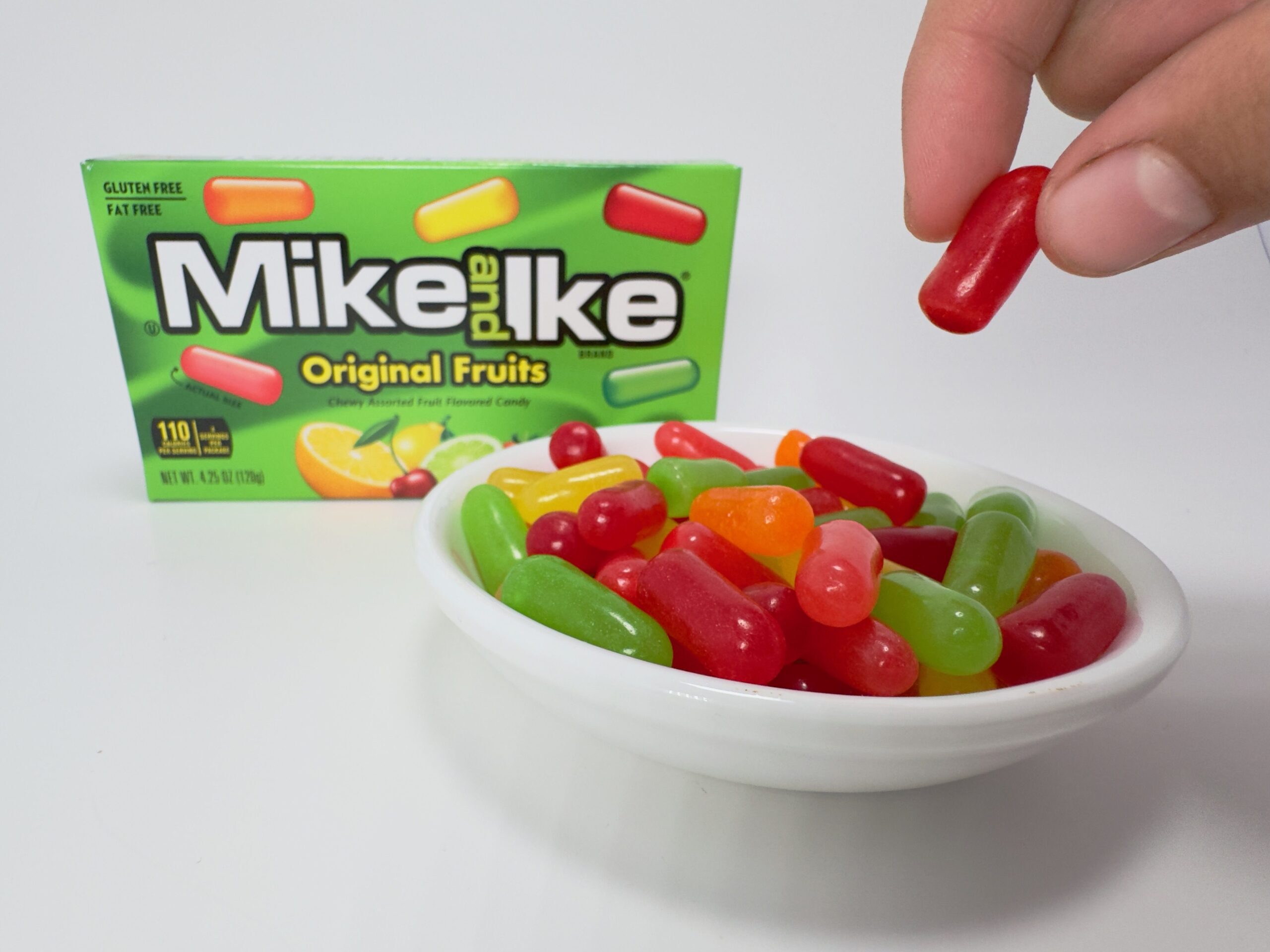 Hand picking up a green Mike and Ike Original Fruits candy from a bowl of mixed cherry, lemon, lime, strawberry, and orange candies with the box in the background.