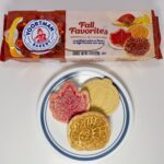 An assortment of Voortman Fall Favorites cookies—a red leaf, a golden turkey, and a yellow pumpkin—on a white plate in front of the product packaging.