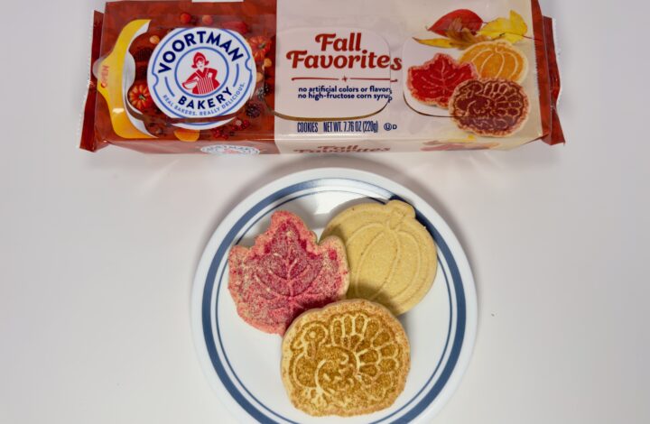 An assortment of Voortman Fall Favorites cookies—a red leaf, a golden turkey, and a yellow pumpkin—on a white plate in front of the product packaging.