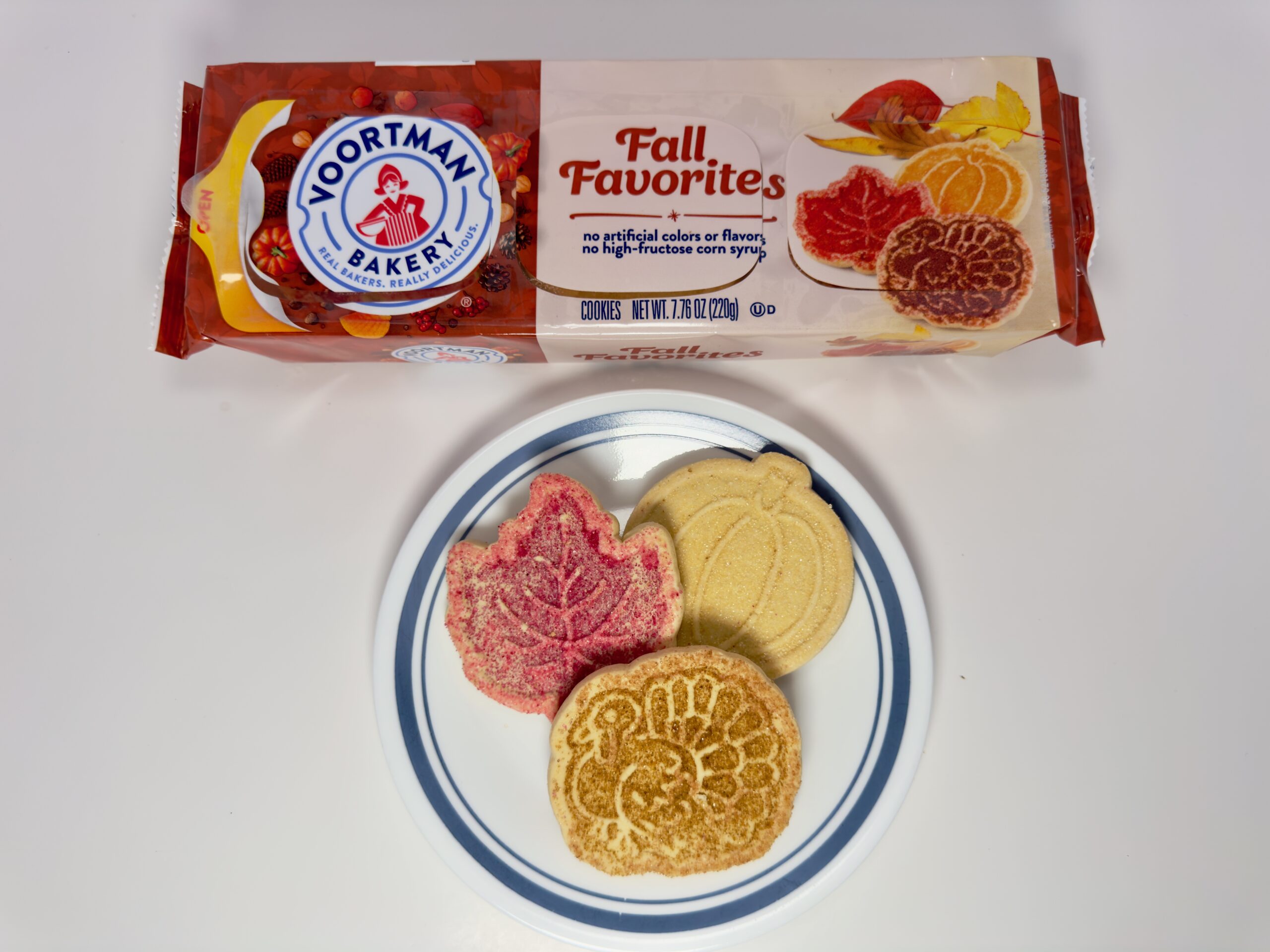 An assortment of Voortman Fall Favorites cookies—a red leaf, a golden turkey, and a yellow pumpkin—on a white plate in front of the product packaging.