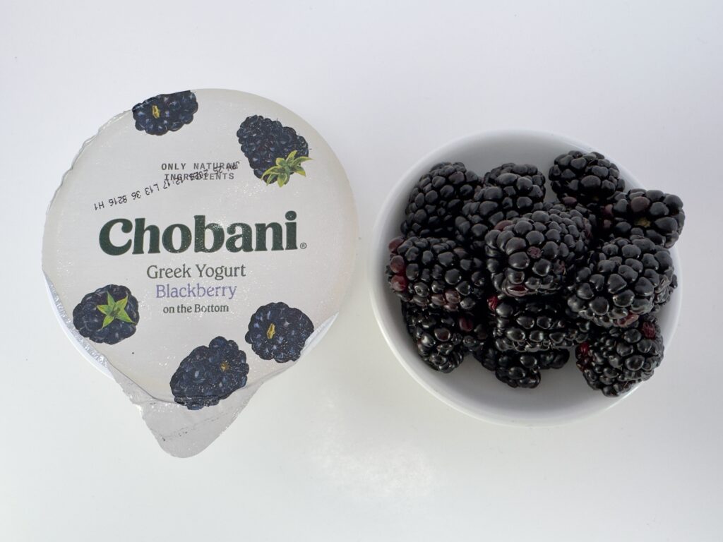 A top-down view of an unopened 5.3 oz cup of Chobani Blackberry Greek Yogurt next to a small white bowl filled with fresh, ripe blackberries.