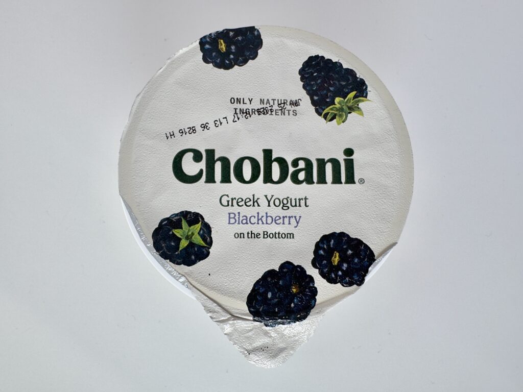 Overhead view of the silver foil lid of a Chobani Blackberry Greek Yogurt cup, showing the brand logo and blackberry illustrations.