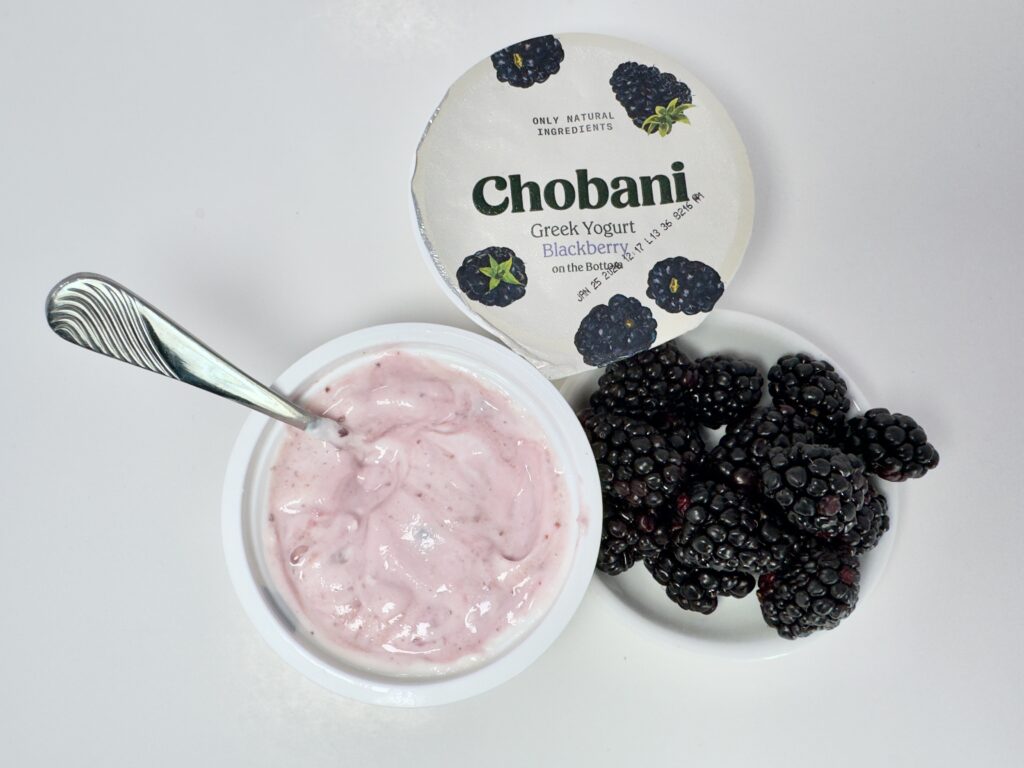 A top view of an open Chobani Blackberry Greek Yogurt cup. A generous pile of fresh, ripe blackberries on a white dish, with an unopened cup in the background.