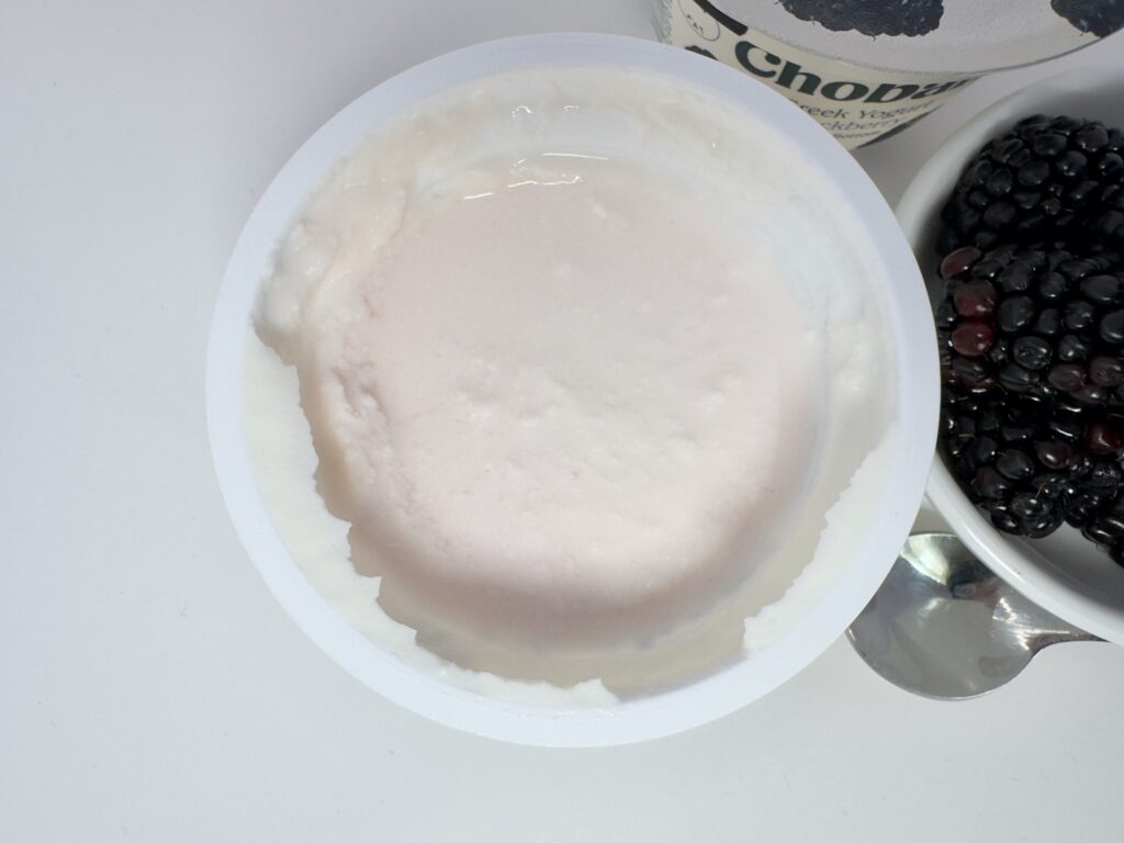 An overhead view of an open cup of Chobani Blackberry on the Bottom Greek yogurt, showing the smooth, unstirred white yogurt surface next to a bowl of fresh blackberries.