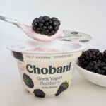 A spoon lifting a fresh, plump blackberry from a cup of Chobani Greek Yogurt Blackberry on the Bottom with a bowl of fresh berries nearby.