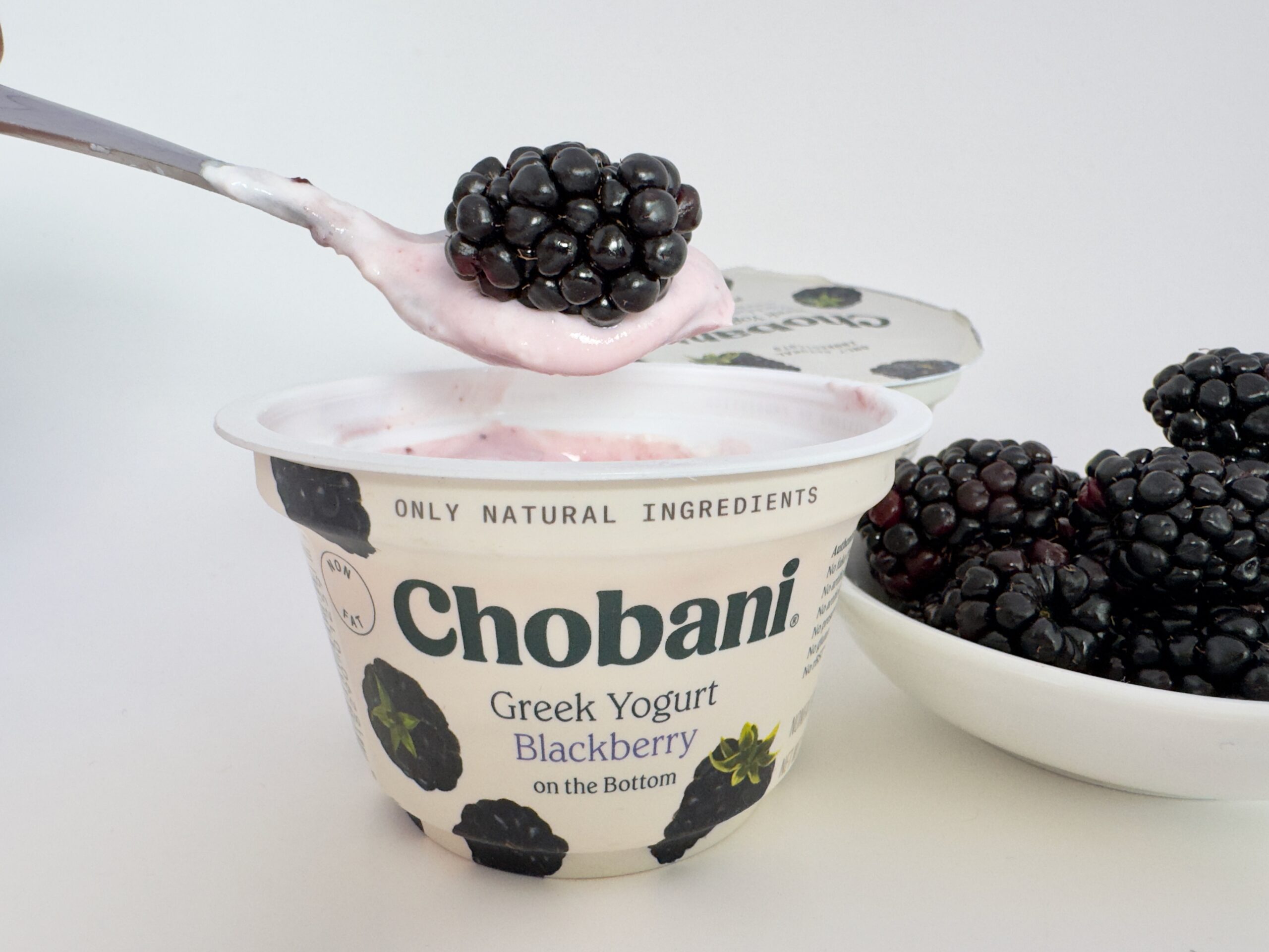 A spoon lifting a fresh, plump blackberry from a cup of Chobani Greek Yogurt Blackberry on the Bottom with a bowl of fresh berries nearby.