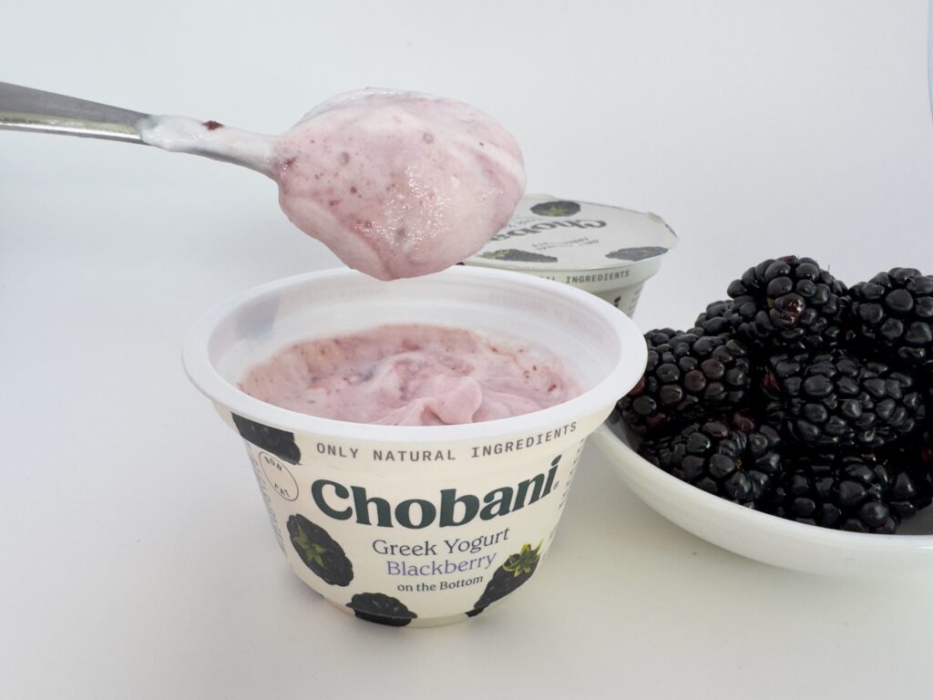 A silver spoon lifting a scoop of mixed Chobani Blackberry Greek Yogurt, showing its thick, creamy pink texture next to a bowl of fresh berries.