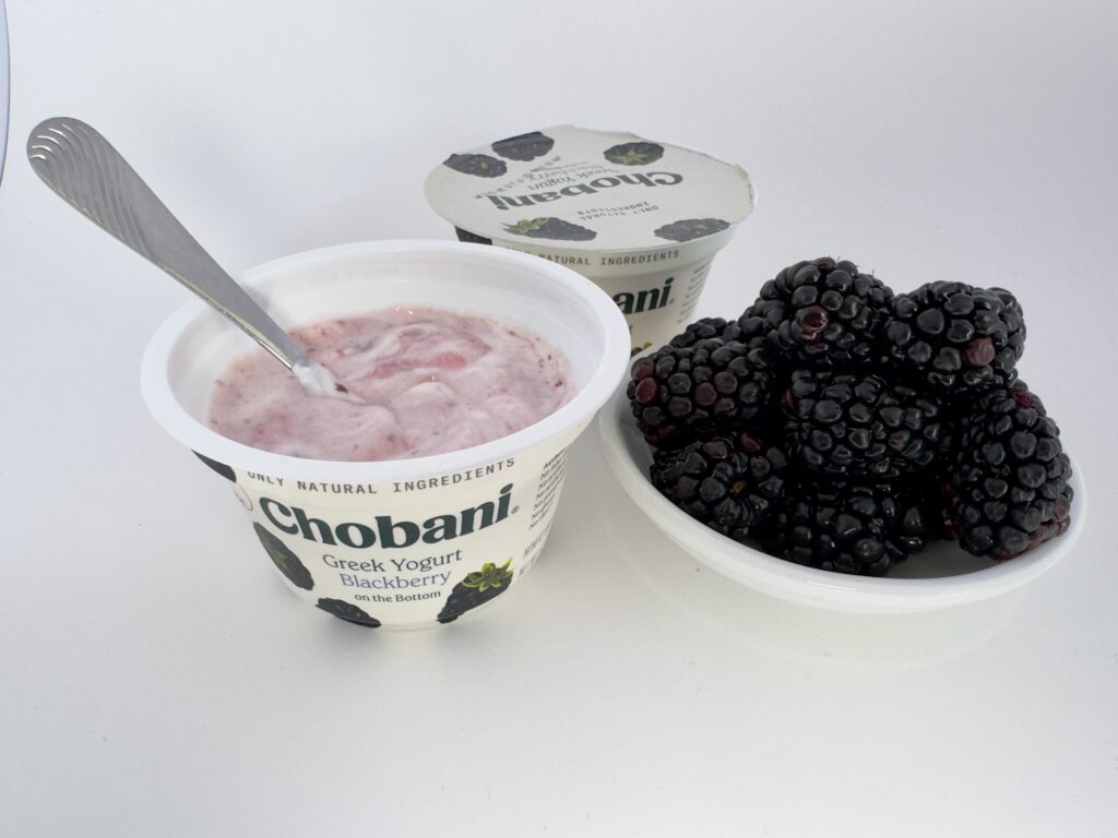 A side-angle view of an open Chobani Blackberry Greek Yogurt cup next to a generous pile of fresh, ripe blackberries, with an unopened cup in the background.