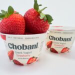 Two cups of Chobani Strawberry on the Bottom Greek Yogurt on a white background, with one cup overflowing with large, fresh strawberries.