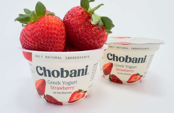 Two cups of Chobani Strawberry on the Bottom Greek Yogurt on a white background, with one cup overflowing with large, fresh strawberries.