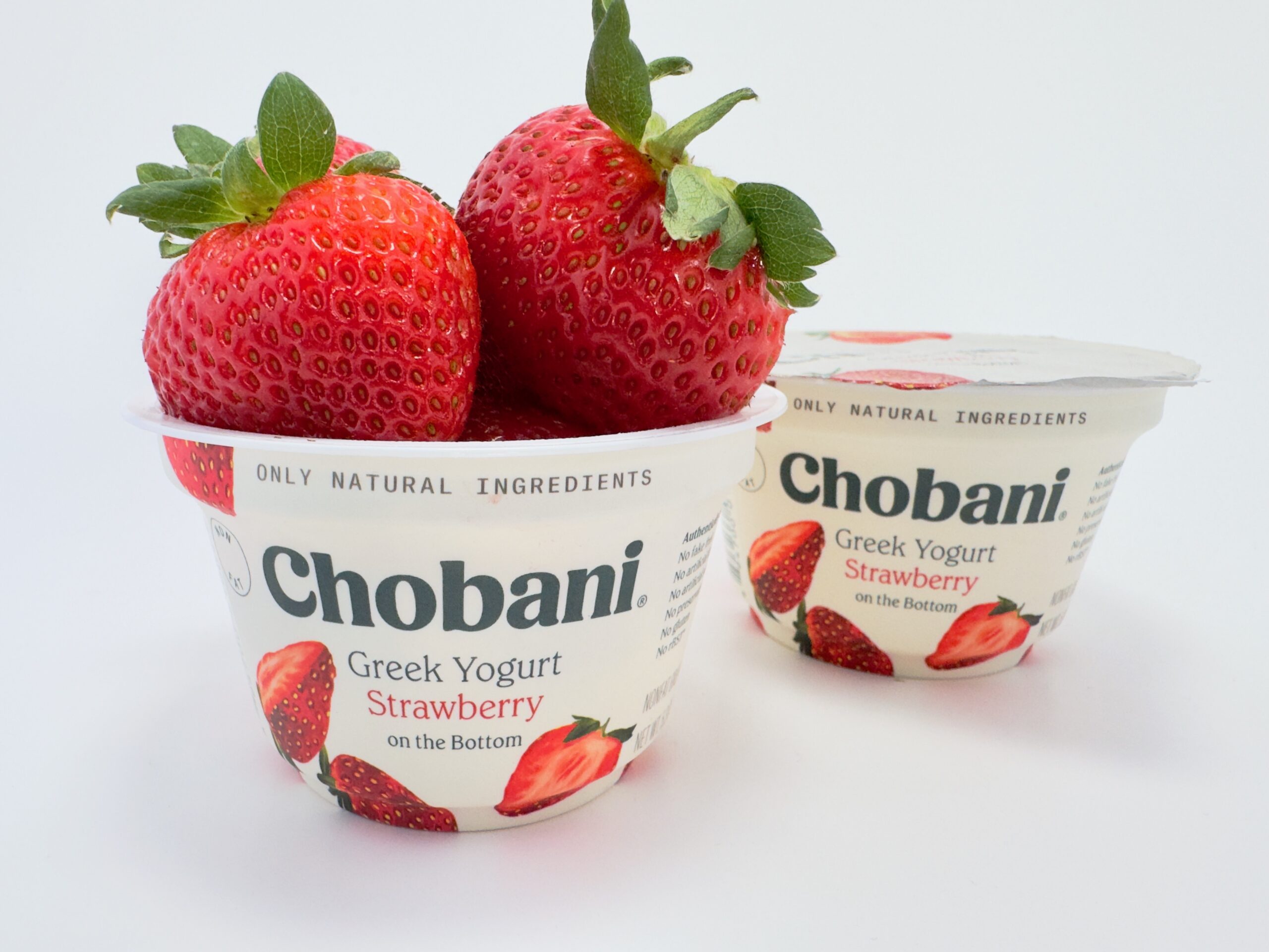 Two cups of Chobani Strawberry on the Bottom Greek Yogurt on a white background, with one cup overflowing with large, fresh strawberries.