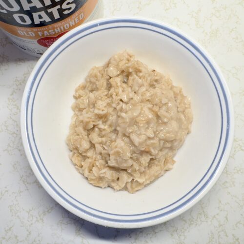 A top-down view of a single serving of cooked, creamy old-fashioned oatmeal in a white bowl with a blue rim, positioned next to a container of Quaker Old Fashioned Oats.
