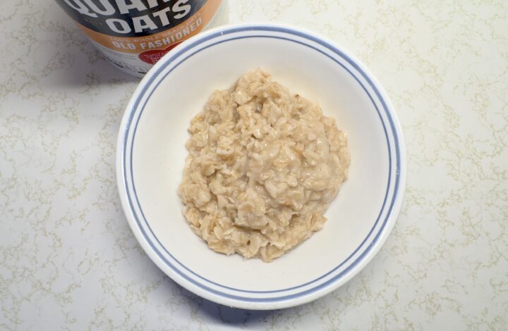 A top-down view of a single serving of cooked, creamy old-fashioned oatmeal in a white bowl with a blue rim, positioned next to a container of Quaker Old Fashioned Oats.