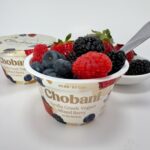 A close-up of a Chobani Hero Vanilla Mixed Berry Greek yogurt cup topped with fresh strawberries, blueberries, raspberries, and blackberries, with a spoon resting inside.