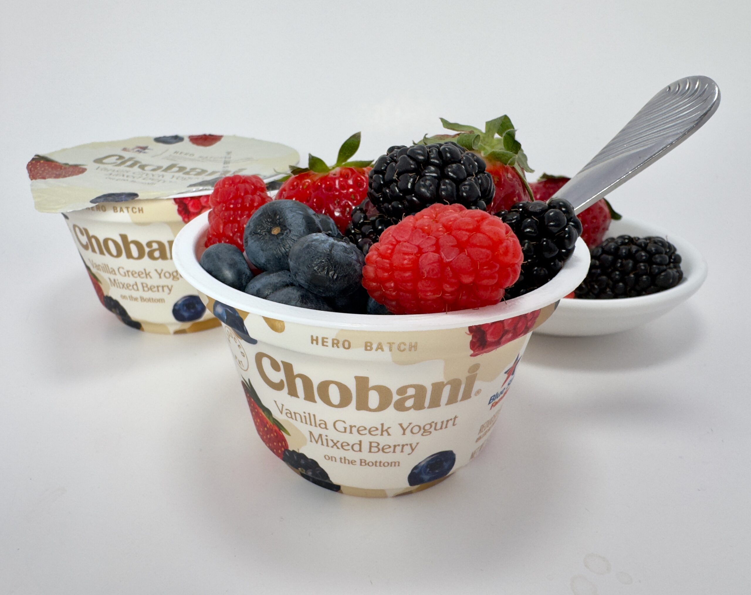 A close-up of a Chobani Hero Vanilla Mixed Berry Greek yogurt cup topped with fresh strawberries, blueberries, raspberries, and blackberries, with a spoon resting inside.