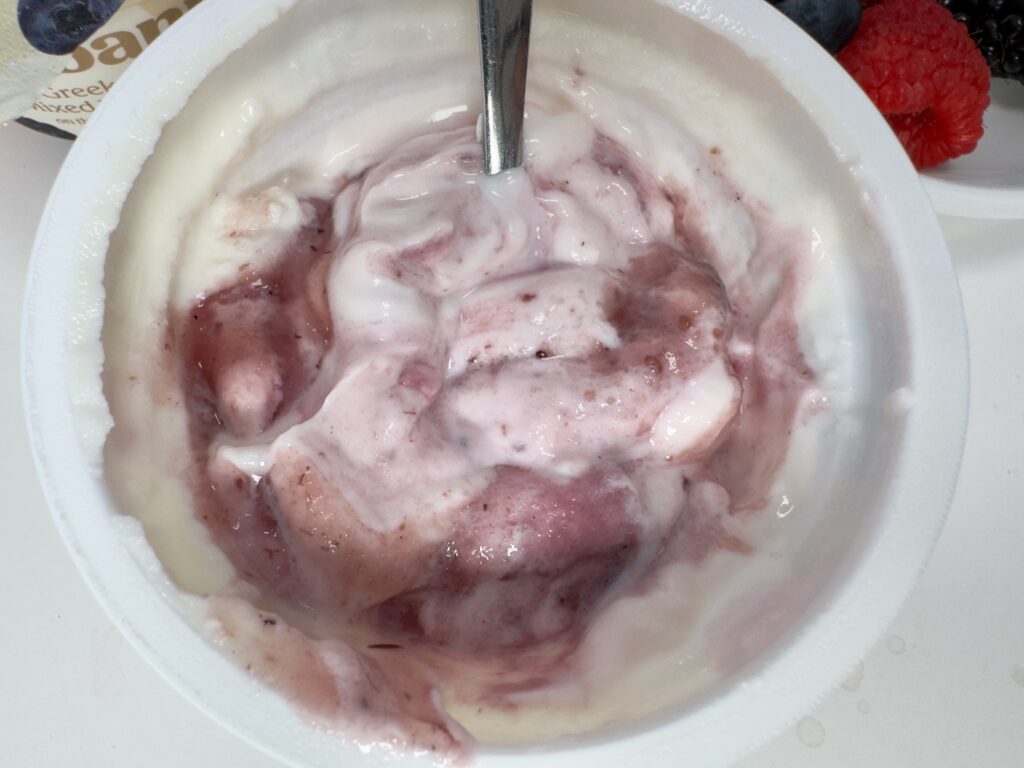 An overhead view of Chobani Vanilla Greek Yogurt partially stirred, showing the purple mixed berry fruit swirls.