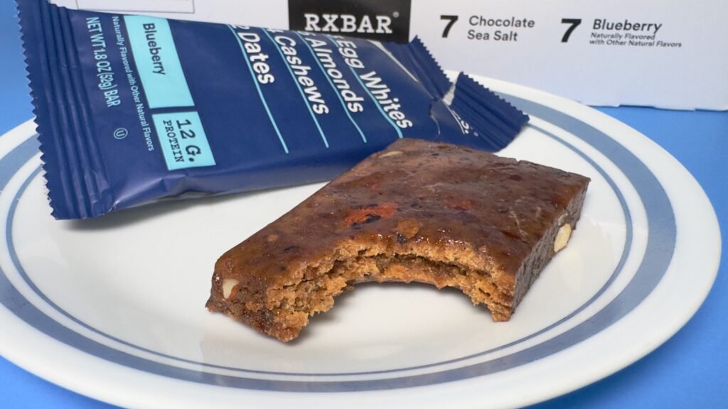 A close-up of a Blueberry RXBAR with a bite taken out, resting on a white plate with its blue wrapper in the background, showing the dense interior and visible fruit pieces.