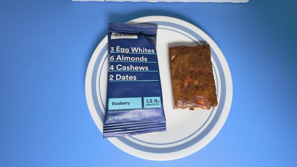 A top-down view of an unwrapped Blueberry RXBAR on a white plate next to its blue wrapper, showing a dense texture with visible nut and fruit pieces.