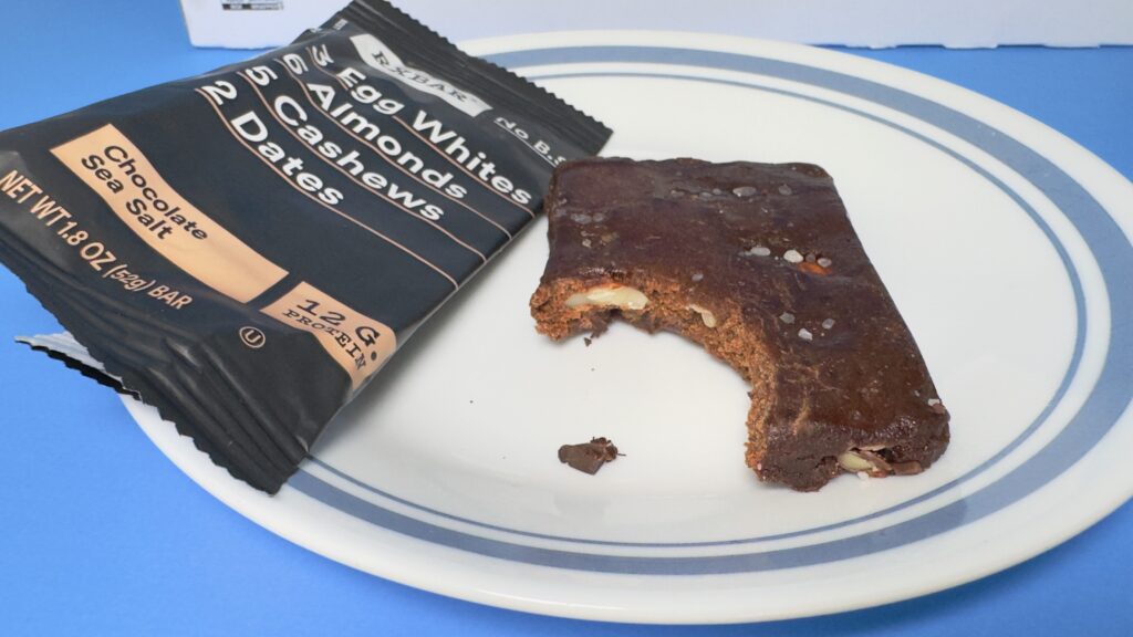 A close-up of a Chocolate Sea Salt RXBAR with a bite taken out, resting on a white plate next to its black wrapper to show the dense interior and visible nut pieces.
