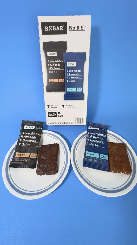 A vertical image of 14-count variety pack of RXBARs from Costco featuring Chocolate Sea Salt and Blueberry flavors, with two unwrapped bars displayed on white plates.